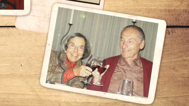 A joyful elderly couple clinks wine glasses, smiling warmly at a dinner table. The woman wears a plaid jacket; the man wears a red cardigan. Nostalgic atmosphere.