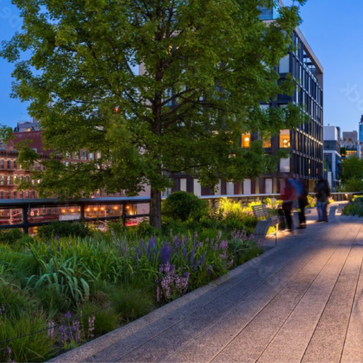 Evening view of the High Line in Manhattan with lit pathways, greenery, and the Empire State Building glowing green.