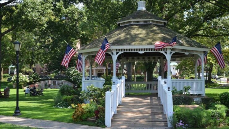 White gazebo in Chappaqua, NY decorated with American flags, surrounded by lush lawns, flowers, and arboreal foliage.