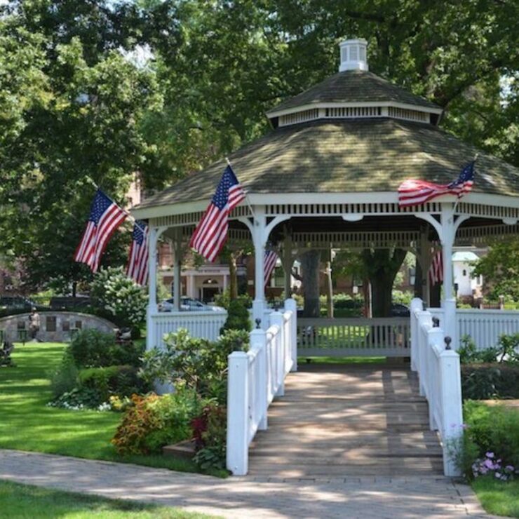 White gazebo in Chappaqua, NY decorated with American flags, surrounded by lush lawns, flowers, and arboreal foliage.