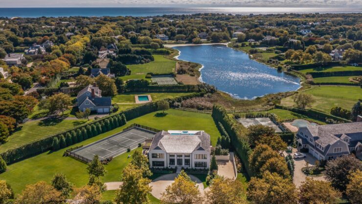 Aerial view of a Hamptons estate with tennis courts, pool, and a lake, near the Atlantic coastline.