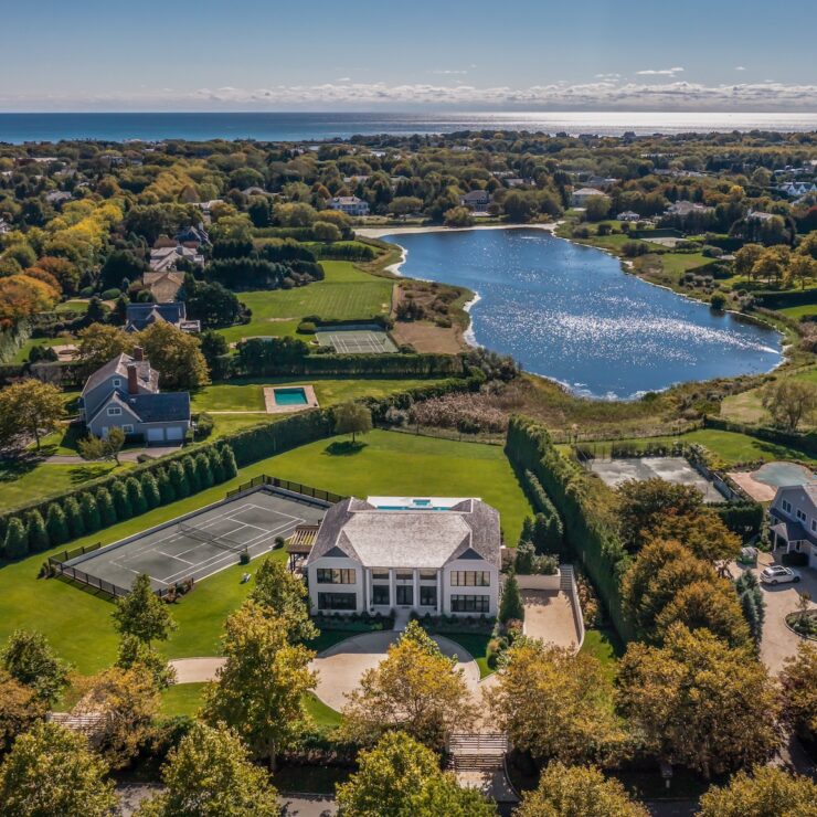 Aerial view of a Hamptons estate with tennis courts, pool, and a lake, near the Atlantic coastline.