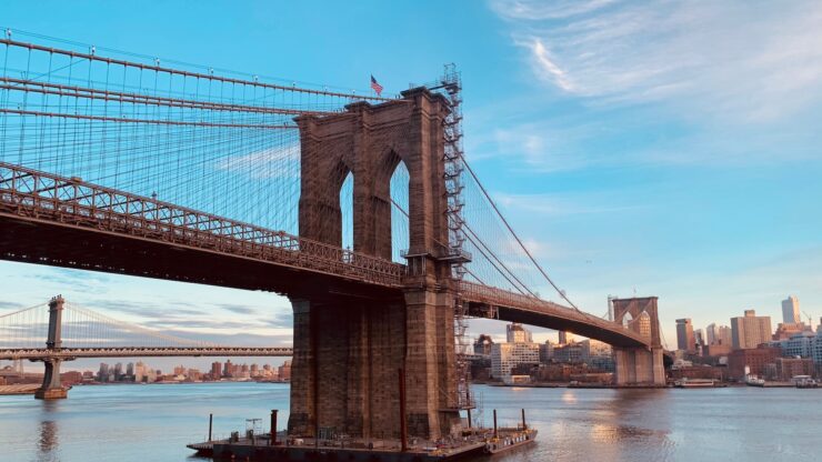 Brooklyn Bridge at sunrise with Manhattan skyline and East River visible under a clear blue sky.