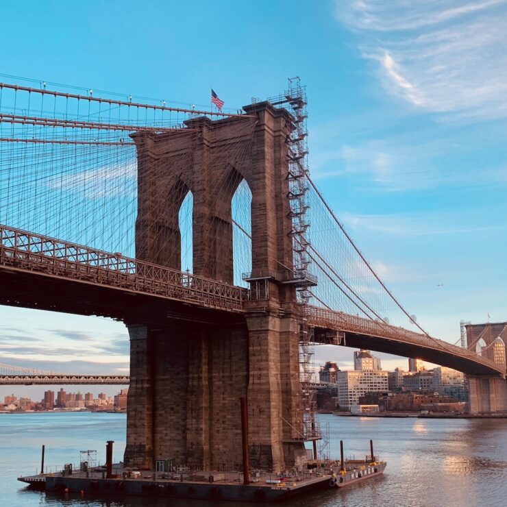 Brooklyn Bridge at sunrise with Manhattan skyline and East River visible under a clear blue sky.