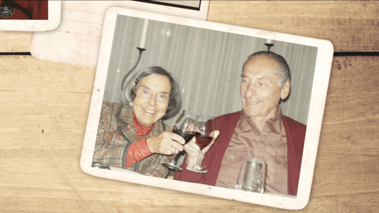 A joyful elderly couple clinks wine glasses, smiling warmly at a dinner table. The woman wears a plaid jacket; the man wears a red cardigan. Nostalgic atmosphere.