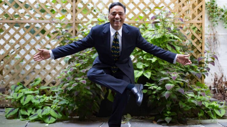 Man in a suit joyfully poses with arms outstretched and one leg crossed, set against a lush garden backdrop with lattice and leafy plants.