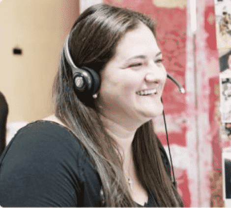 Smiling woman with long brown hair wearing a headset, seated indoors. The background is red and white.