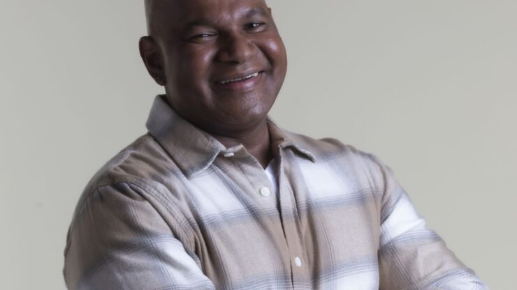 A smiling man is wearing a plaid shirt against a plain background.