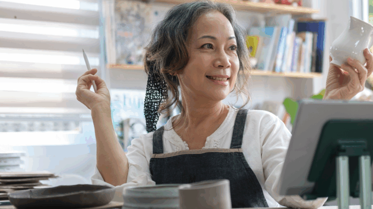Woman in an apron sitting at a table, holding a white ceramic piece and a tool, smiling. Bookshelves with books in the background.