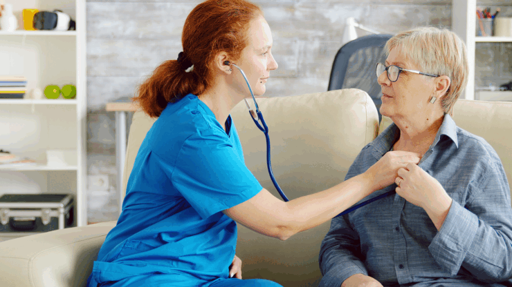 Nurse in blue scrubs listens to an elder woman’s heartbeat with a stethoscope on a beige couch.