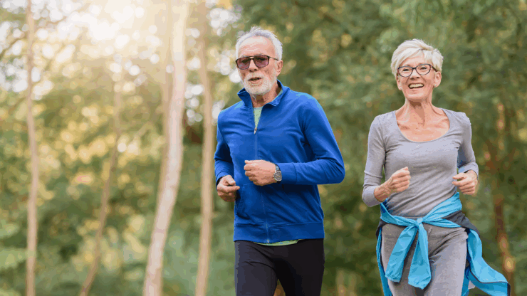 Elderly man in blue jacket and woman in gray, both jogging joyfully in a sunlit forest.
