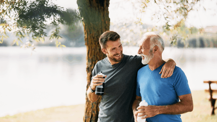 A smiling older man and younger man with water bottles embrace under a tree by a lake.