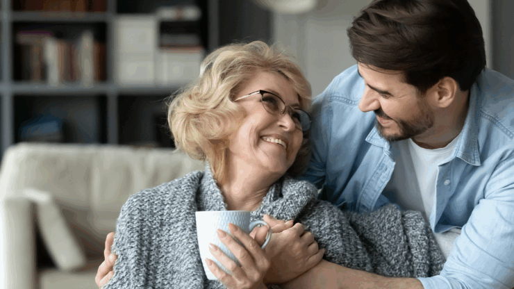 A smiling older woman in glasses, wrapped in a gray blanket, holds a mug while a younger man embraces her.