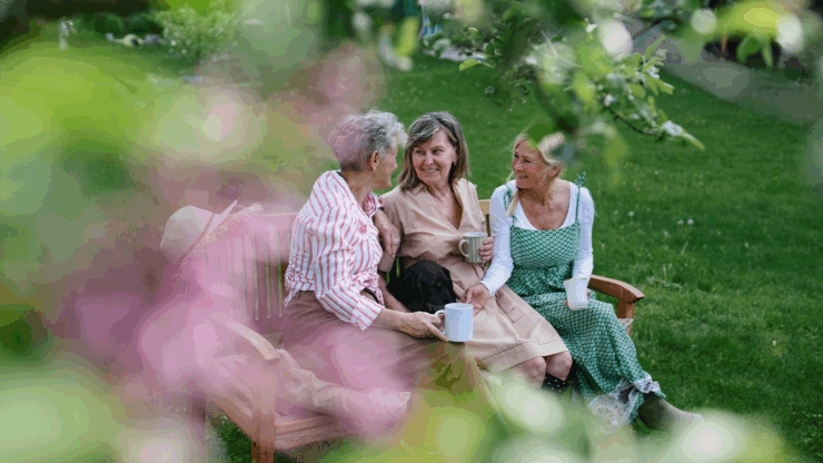 Three women sit on a wooden bench in a lush garden, smiling and conversing with mugs in hand. The scene is framed by soft pink blossoms.