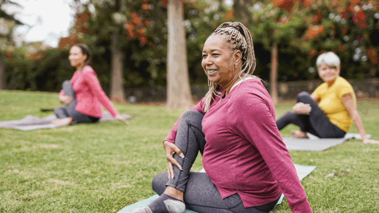 Three women practice yoga in a park. The woman in the foreground smiles while doing a seated twist.
