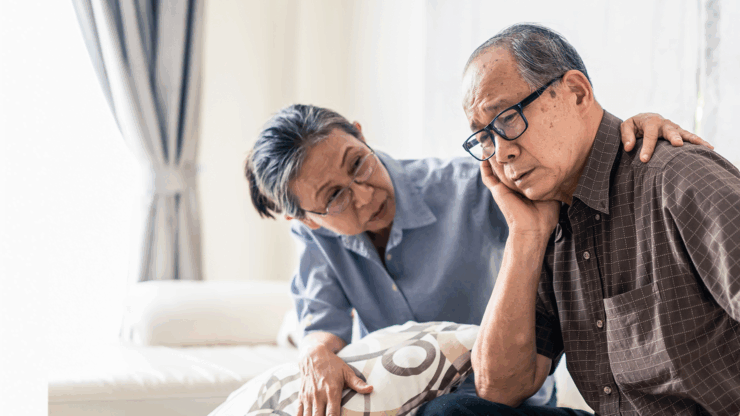 An elderly couple sits in a bright living room, with the woman comforting the man, who looks worried.