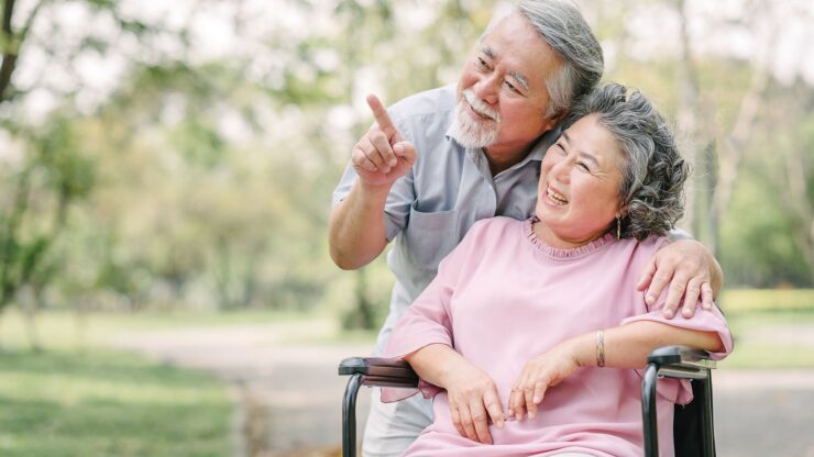 An elderly man stands beside a smiling woman in a wheelchair, pointing at something in the distance. They are in a park.