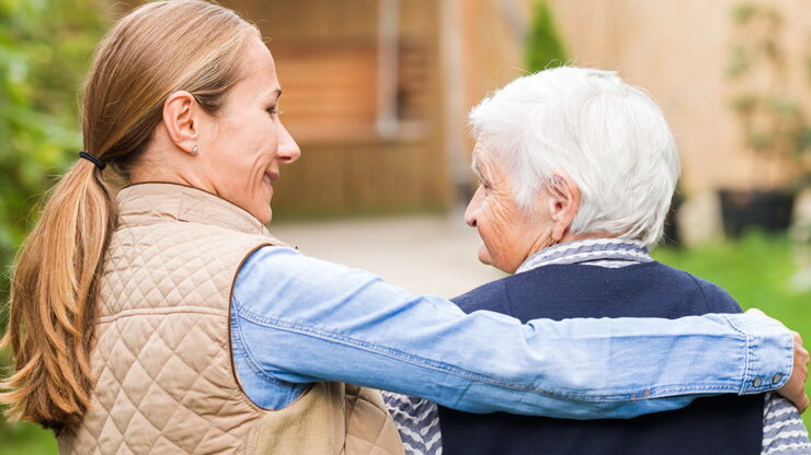 A woman with a ponytail and beige vest gently embraces an elderly woman with short white hair.