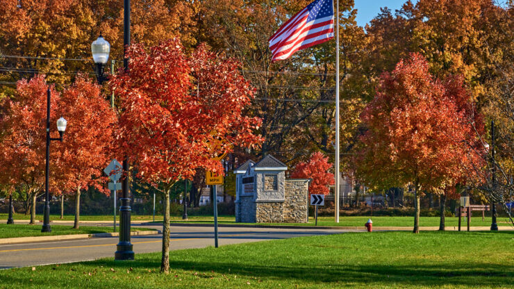 Autumn scene with vivid red and orange trees lining a street beneath a clear blue sky. An American flag flutters prominently in the foreground.
