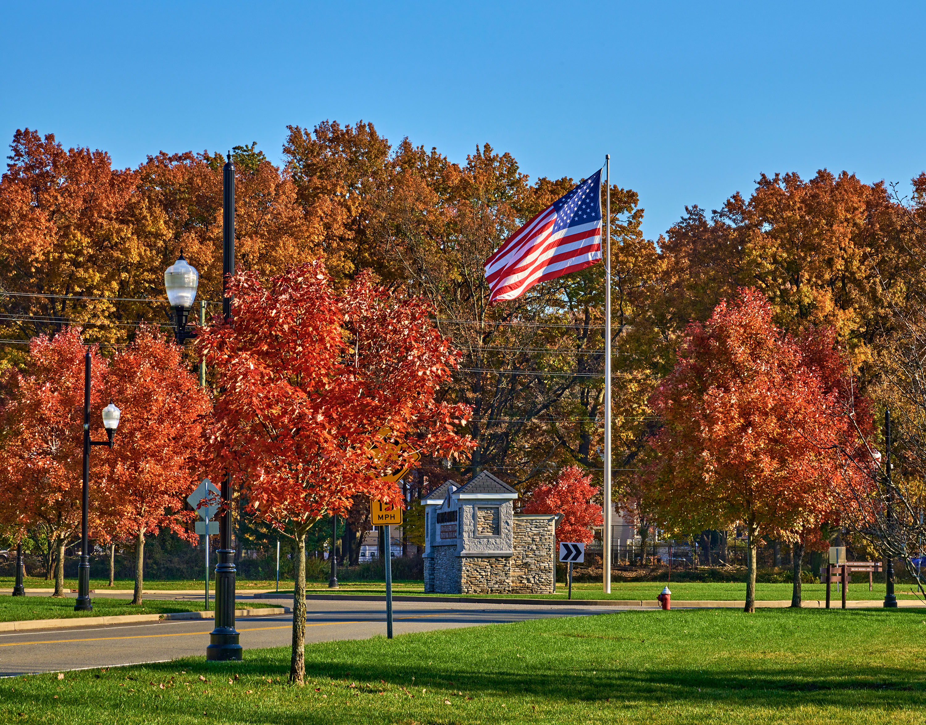 Autumn scene with vivid red and orange trees lining a street beneath a clear blue sky. An American flag flutters prominently in the foreground.