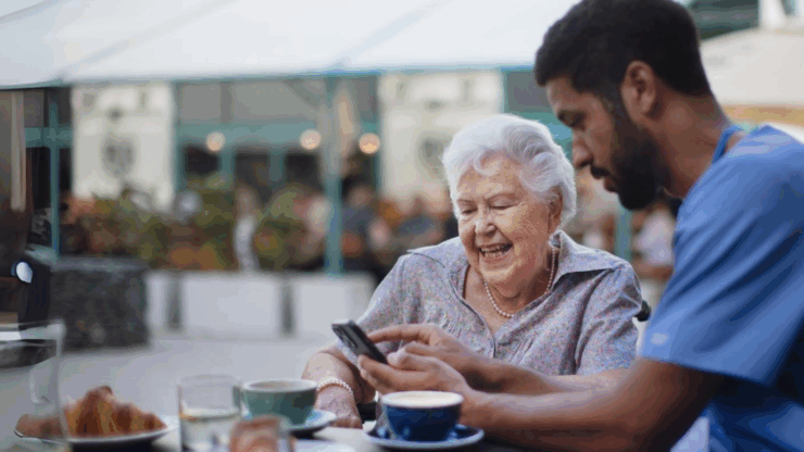 Male caregiver assisting a smiling elder woman with her phone at a cafe table outside