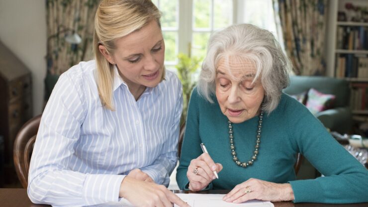 A woman in a striped shirt assists an older woman in a teal sweater with paperwork.