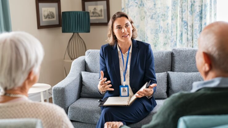 A professional woman in a blue suit consults with an elderly couple on a sofa, holding a notepad.