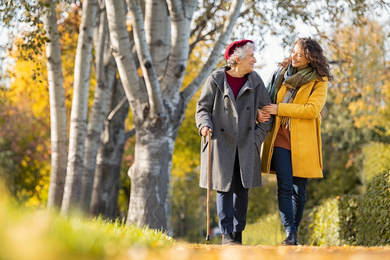 Older adult walking with caregiver on a fall day, both smiling and warmly dressed, surrounded by birch trees.