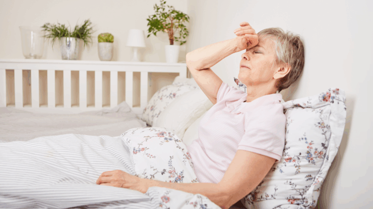 An elderly woman in a pink shirt rests against pillows in bed, looking fatigued, with her hand on her forehead.