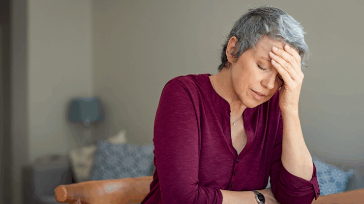A woman with short gray hair wearing a maroon shirt sits with her hand on her forehead, looking stressed