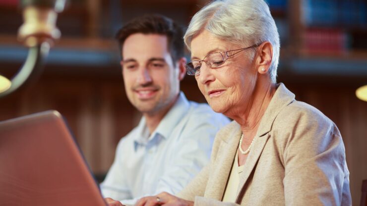 An elderly woman in glasses types on a laptop, smiling warmly. A younger man, also smiling, sits beside her.