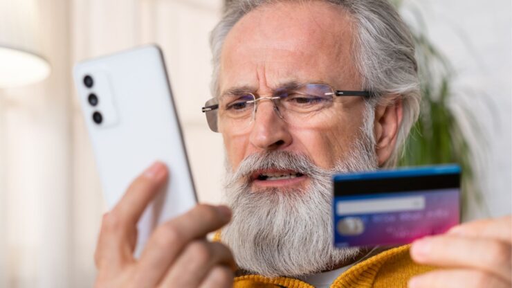 Elderly man with glasses, wearing a yellow sweater, looks confused while holding a smartphone and a credit card.