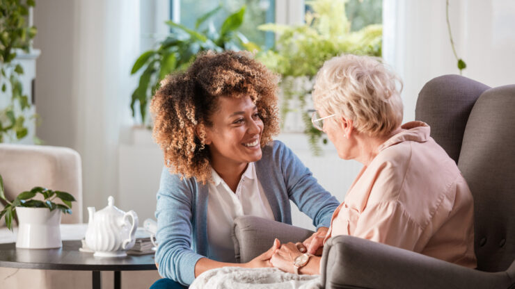 A young woman in a blue sweater holds hands with an older woman in glasses, sitting close on an armchair, sharing an emotional and caring moment.