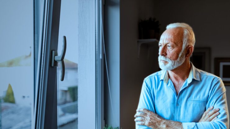 Elderly man with white beard in blue shirt gazes thoughtfully out of an open window, arms crossed.