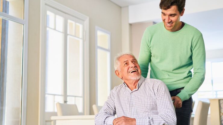 A smiling elderly man in a wheelchair is joyfully interacting with a young man standing behind him in a bright, sunlit room.