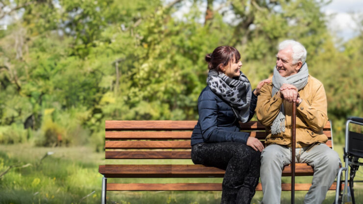 A young woman and an elderly man are sitting on a park bench, engaging in a friendly conversation with greenery in the background.