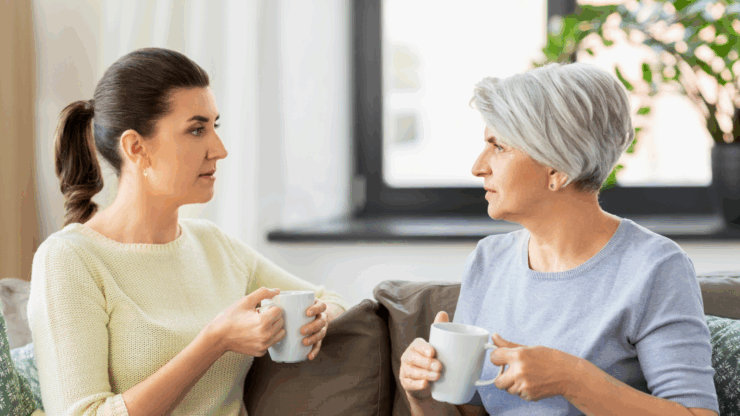 Two women sitting on a couch, facing each other, holding white mugs. One has dark hair in a ponytail, and the other has short gray hair. They appear engaged in conversation.