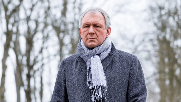 Elderly man in a gray coat and scarf stands outdoors on an overcast day, with bare tree branches in the blurred background, looking serious.