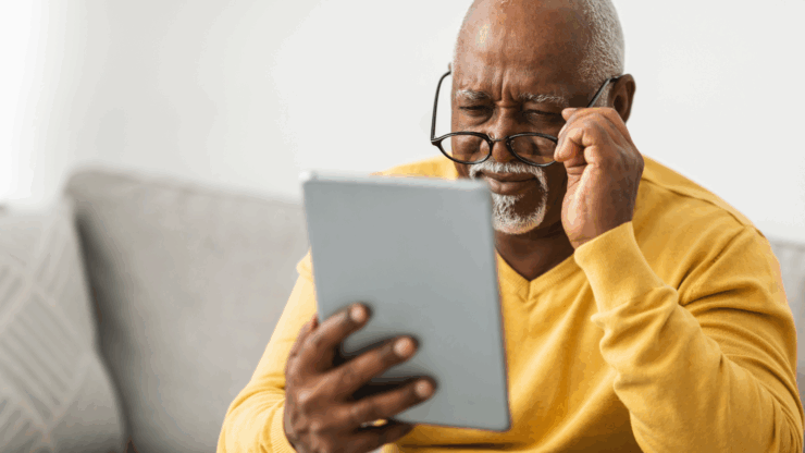 Elderly man with glasses squints at a tablet, wearing a yellow sweater, sitting on a sofa.
