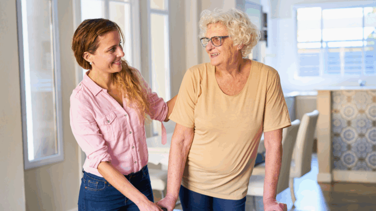 A smiling woman in a pink shirt provides support to an elderly woman using a walker.
