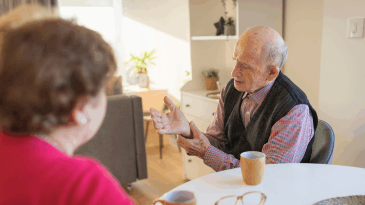 Elderly man and woman at a table with mugs, engaged in a conversation.