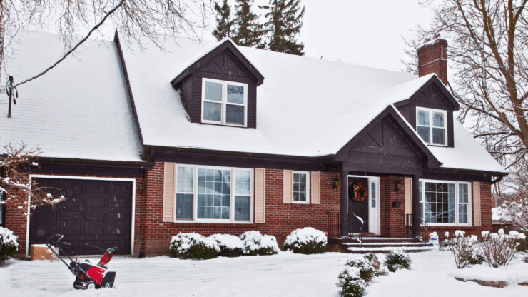 Snow-covered brick house with Christmas wreath on the door, surrounded by snowy trees and bushes. A red snow blower sits on the driveway.