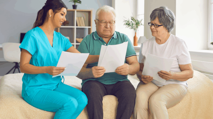 A nurse in blue scrubs smiling while discussing documents with an elderly couple seated on a bed.