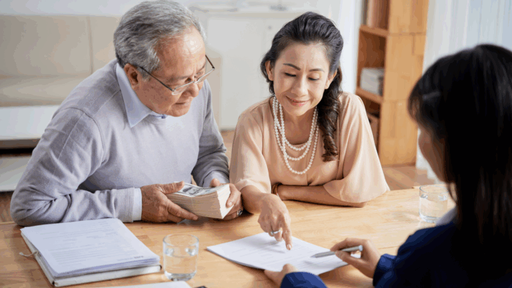 Elderly couple reviews documents with a financial advisor at a table. The woman points at a paper, while the man holds a stack of cash, conveying focus and attention.