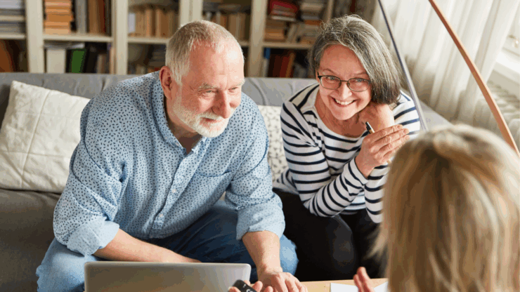 Elderly couple smiling, sitting on a sofa with a laptop, discussing colorful charts with a woman holding a calculator.