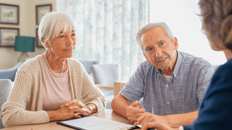 An elderly couple sits at a table in discussion with a professional woman. The couple appears attentive and engaged, with documents and a pen on the table.