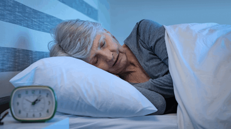 A serene elderly woman with gray hair sleeps peacefully in bed, under a white blanket. An alarm clock on a nightstand shows early morning.