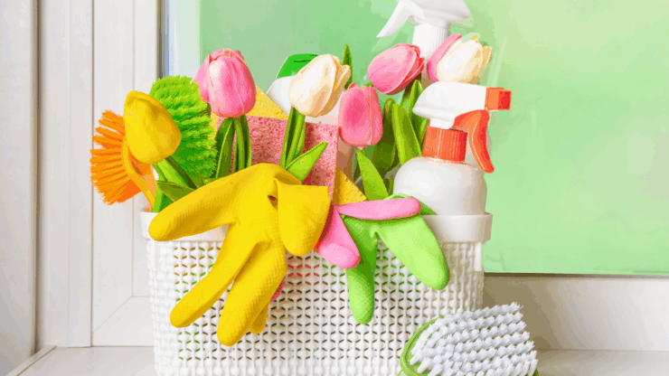 A basket of cleaning supplies with colorful brushes, rubber gloves, spray bottles, and sponges, accented by pink and yellow tulips, sits by a window.