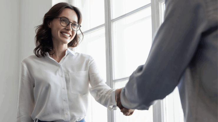Smiling woman with glasses, wearing a white shirt and jeans, shakes hands with a person in a bright room with large windows