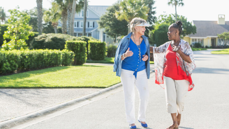 Two women walk and chat on a sunny street lined with greenery and palm trees. One wears a blue outfit, the other red.